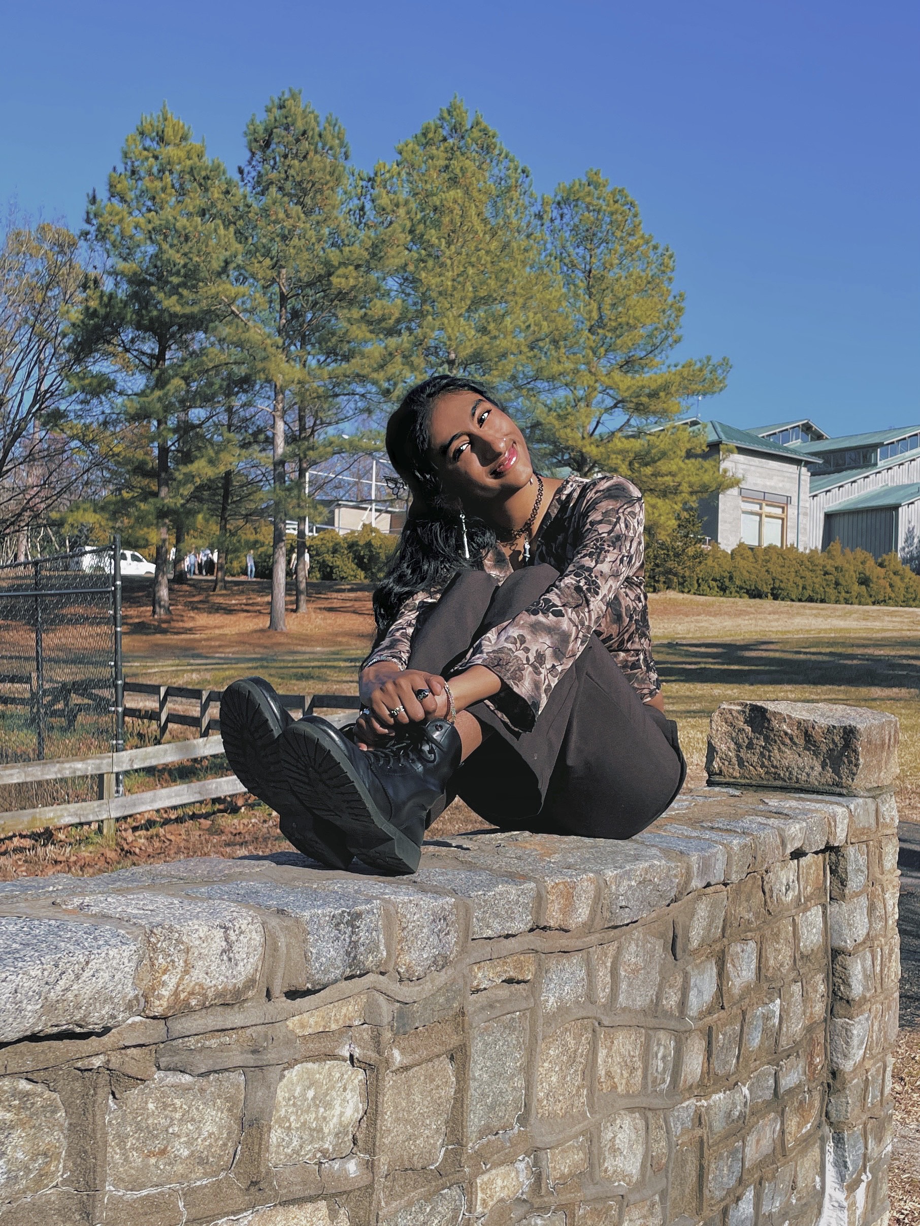 Arya sitting on a stonewall wearng black jeans and a dark top.